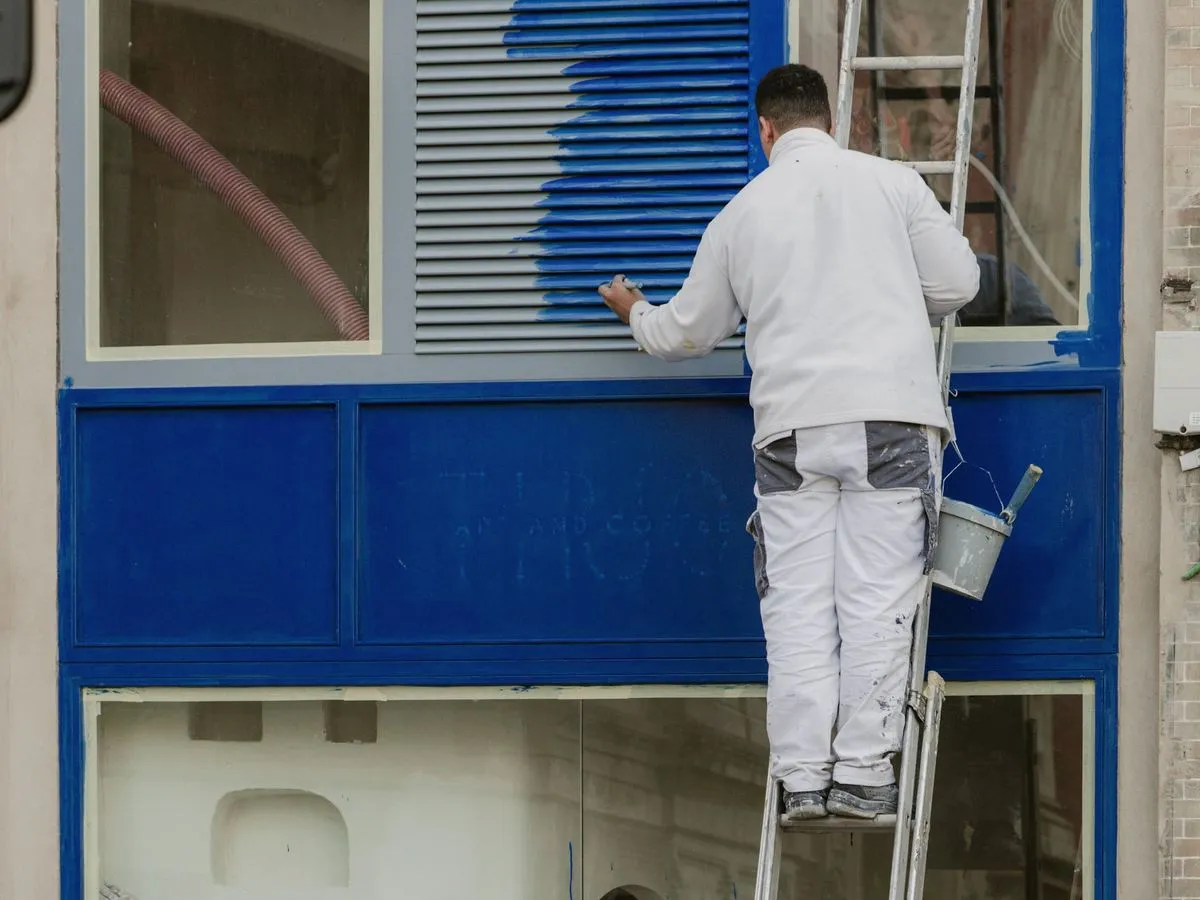 Maler im weissen Arbeitsanzug streicht Fensterläden auf einer Leiter